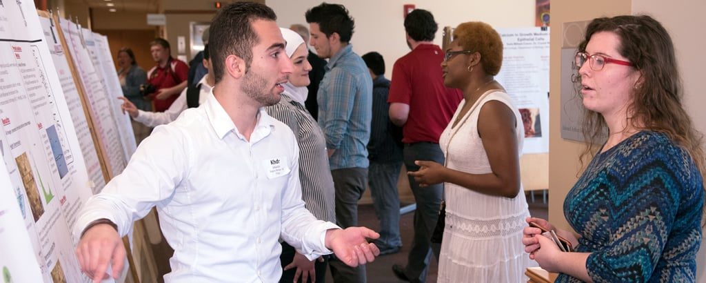 A student presents his poster research to other students nearby.